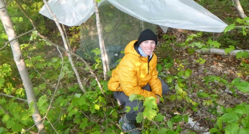 A person smiles beside their tarp shelter set up in a wooded area